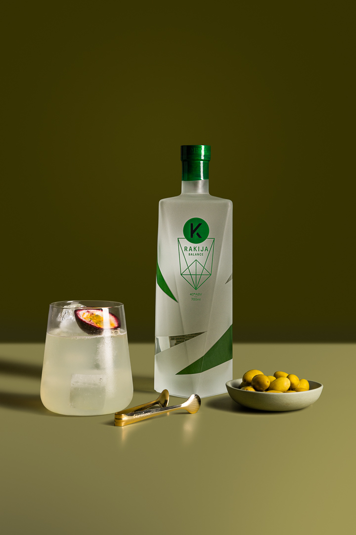 Frosted Kabina rakija Balance bottle sitting in an olive green room, with full glass garnished with half a passionfruit, small tongs, and a bowl of olives.