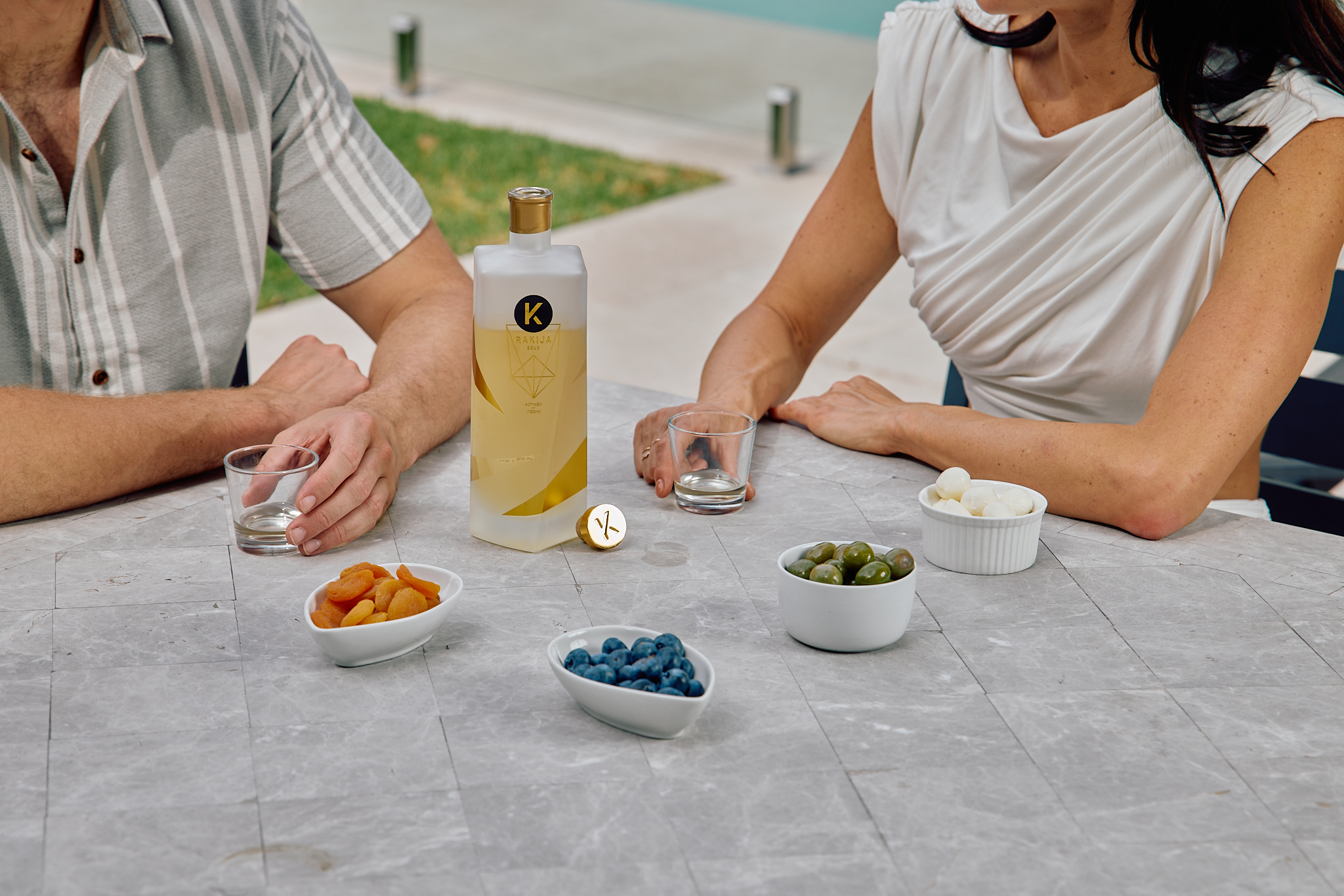 Man and woman sitting at a tiled table with a frosted bottle of Kabina gold rakija, holding empty glasses and surrounded by small bowls filled with dried apricots, blueberries, olives and pickled onions.