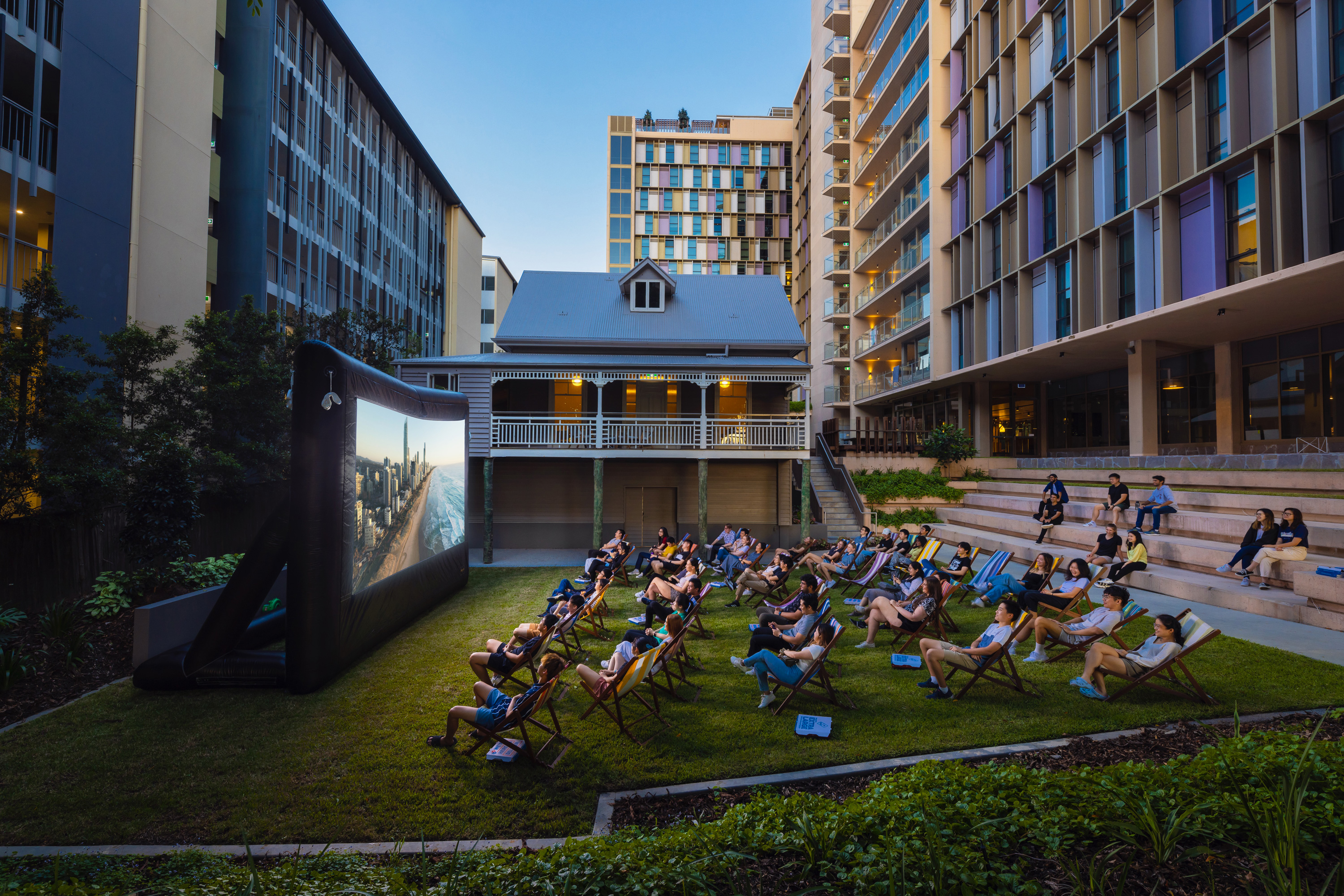 A large group of students sit outdoors in deck chairs facing an inflatable screen for an outdoor cinema at the University of Queensland.