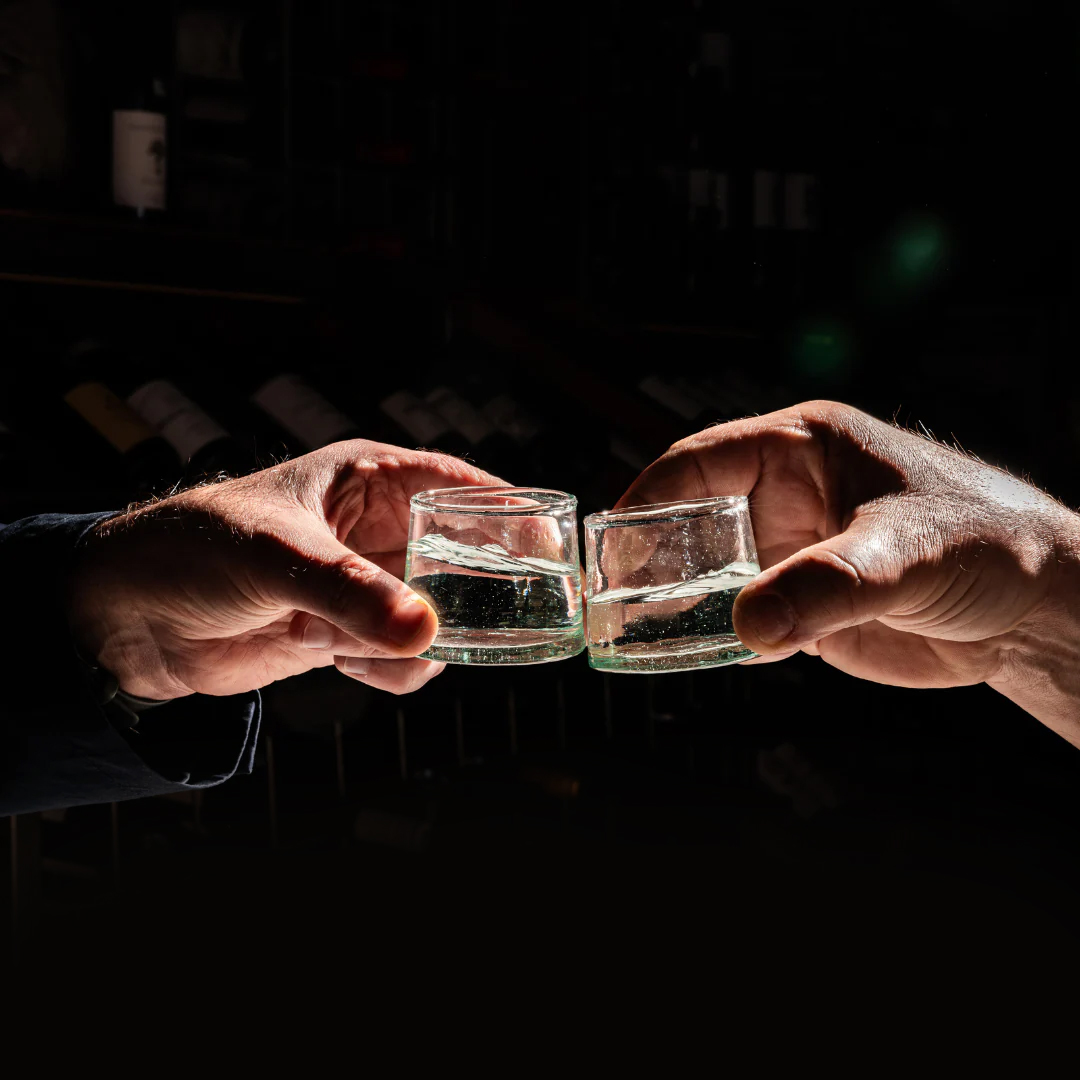 Two hands clinking together small shots of rakija in a dark setting.