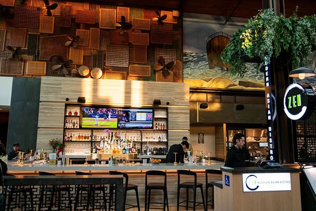 Modern Asian bistro bar interior with wooden stools, two TV screens, and bartenders behind the counter.