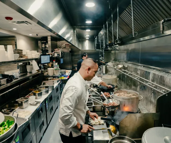 Two chefs cooking in a stainless steel commercial kitchen with pots and pans on the stove.