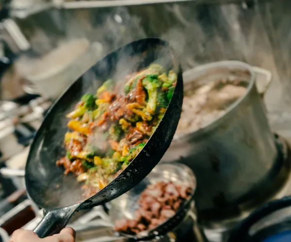 Hand holding a pan tossing stir-fried vegetables over a stove with steam rising and pots in the background.