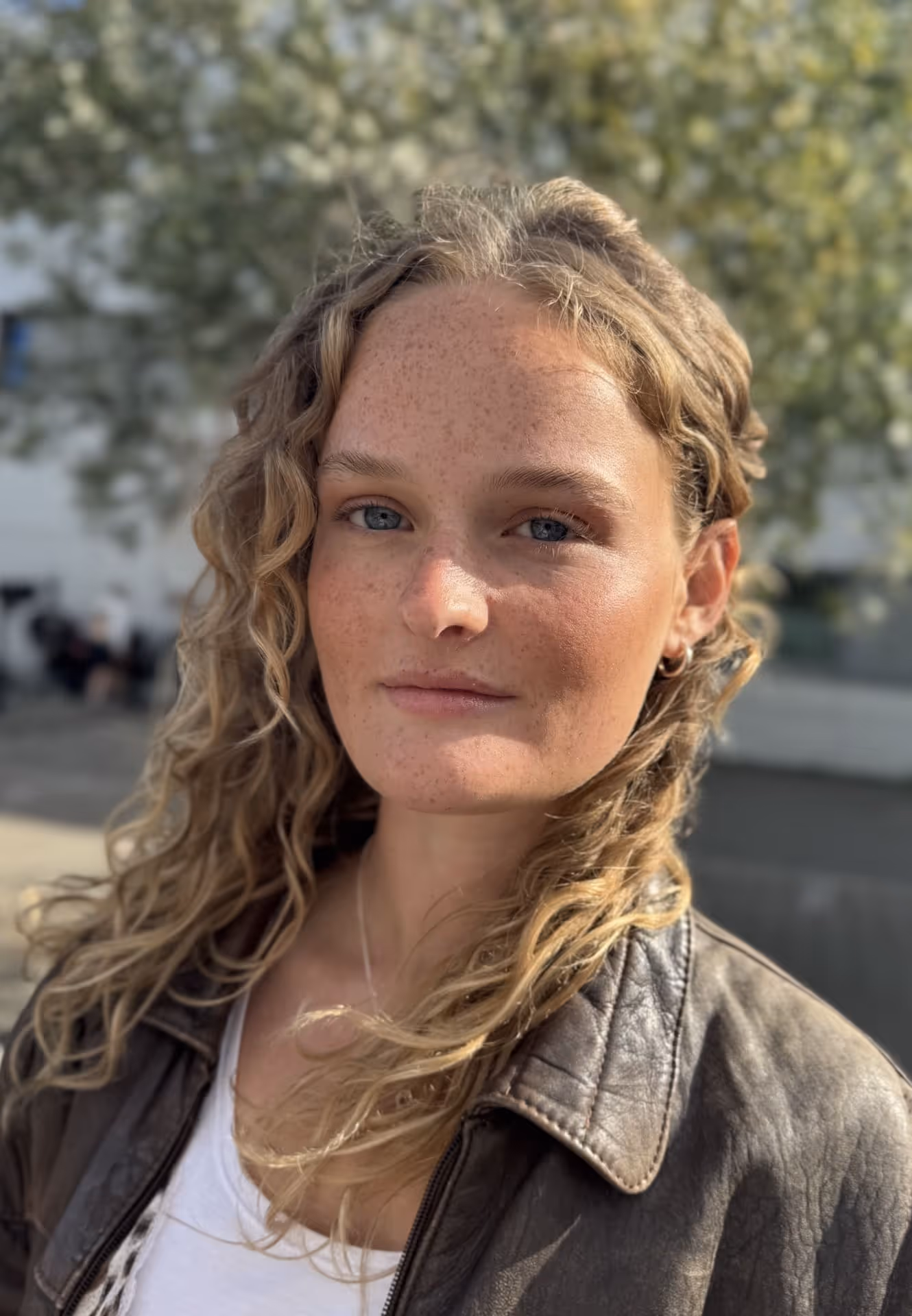 Close-up portrait of a woman with curly blonde hair, freckles, and blue eyes wearing a brown leather jacket outdoors.