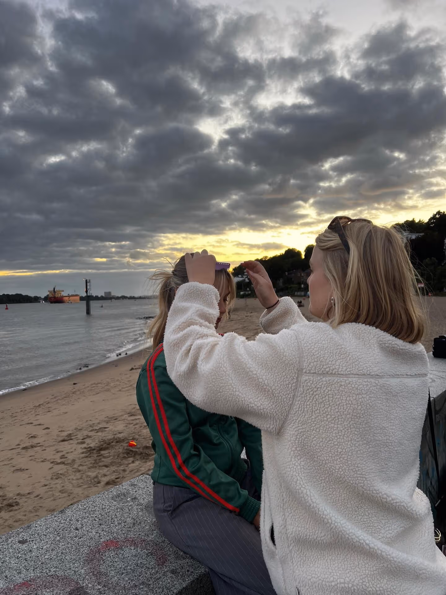Two women on a beach at sunset, one adjusting the other's sunglasses while sitting on a stone ledge near the water.