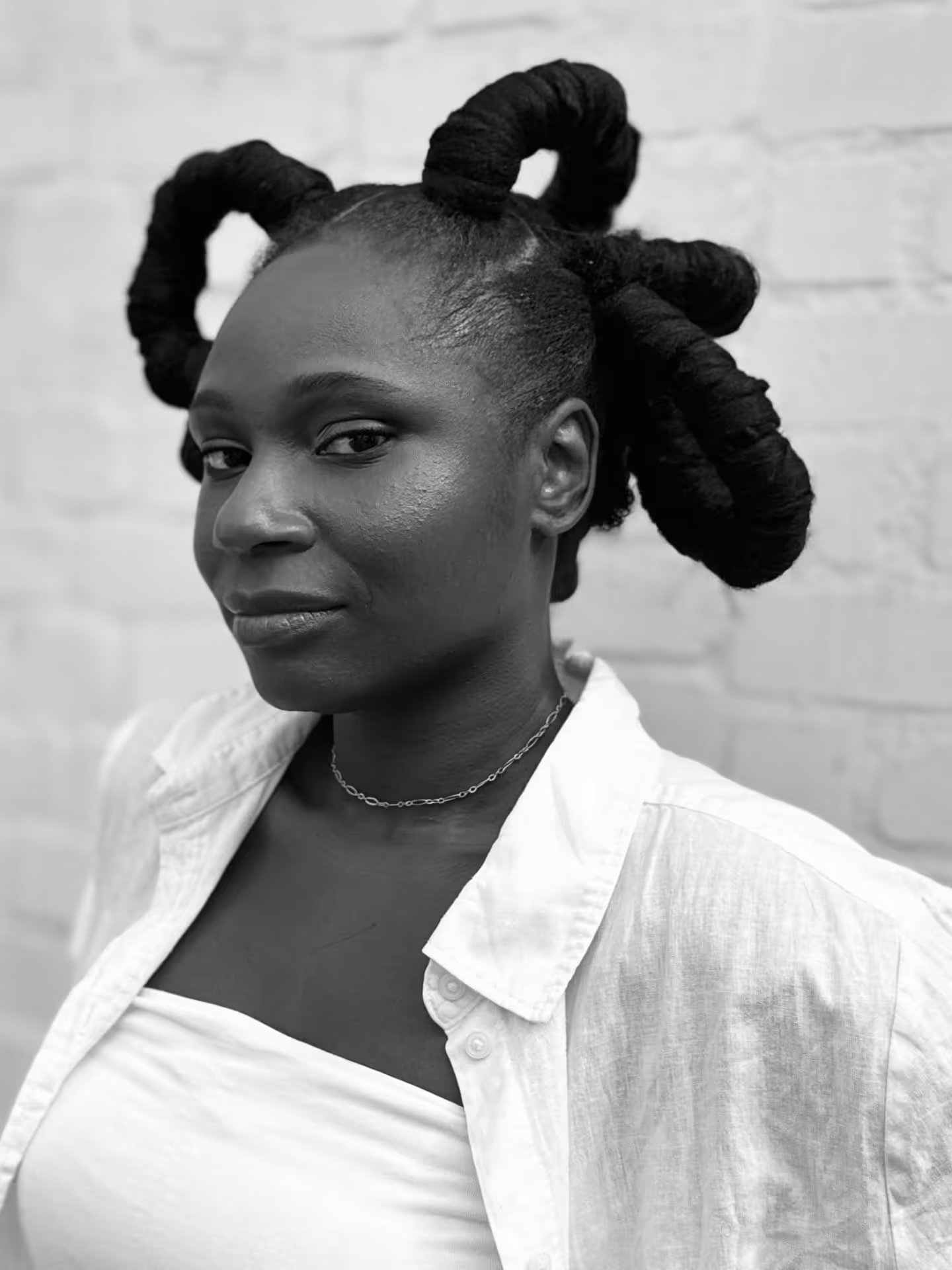 Portrait of a confident woman with artistic twisted hair wearing a white shirt and chain necklace, posing against a brick wall.