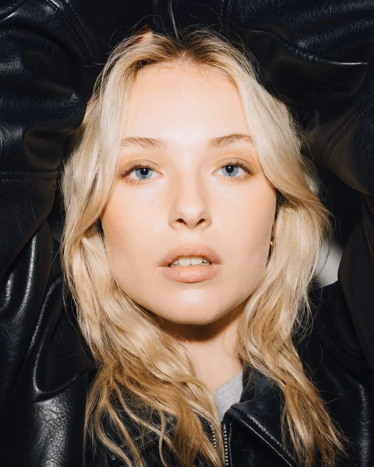 Close-up portrait of a blonde woman with blue eyes wearing a black leather jacket, looking directly at the camera.