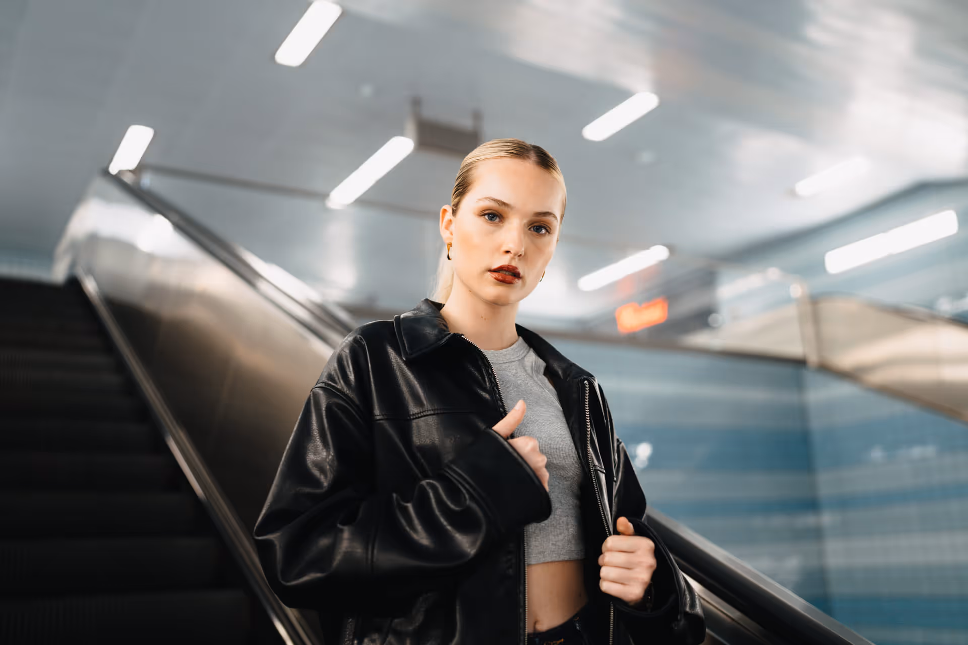 Young woman in a black leather jacket and gray crop top standing near an escalator in a modern subway station.