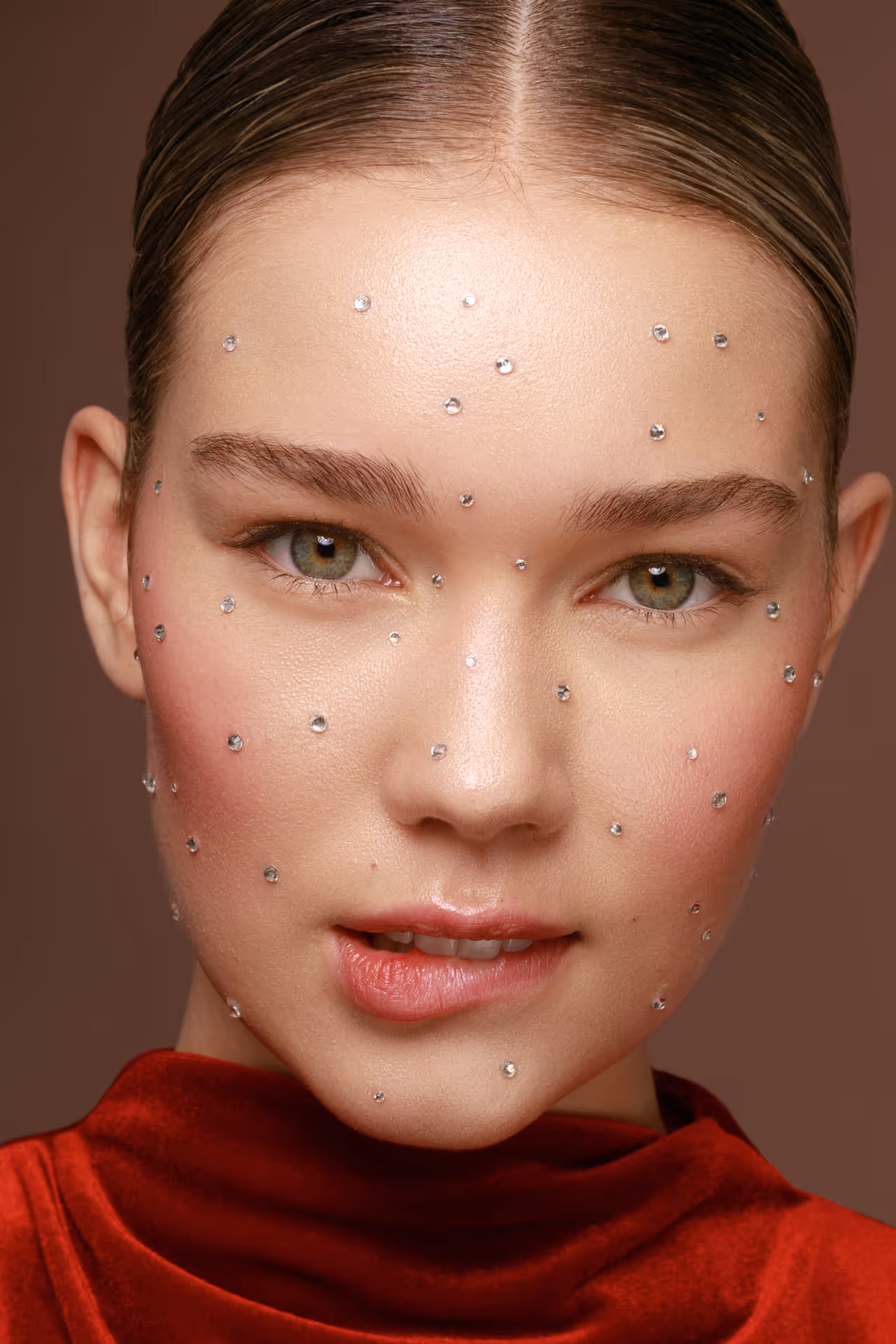 Close-up of a woman with light skin, green eyes, and small rhinestones scattered across her face wearing a red velvet top.