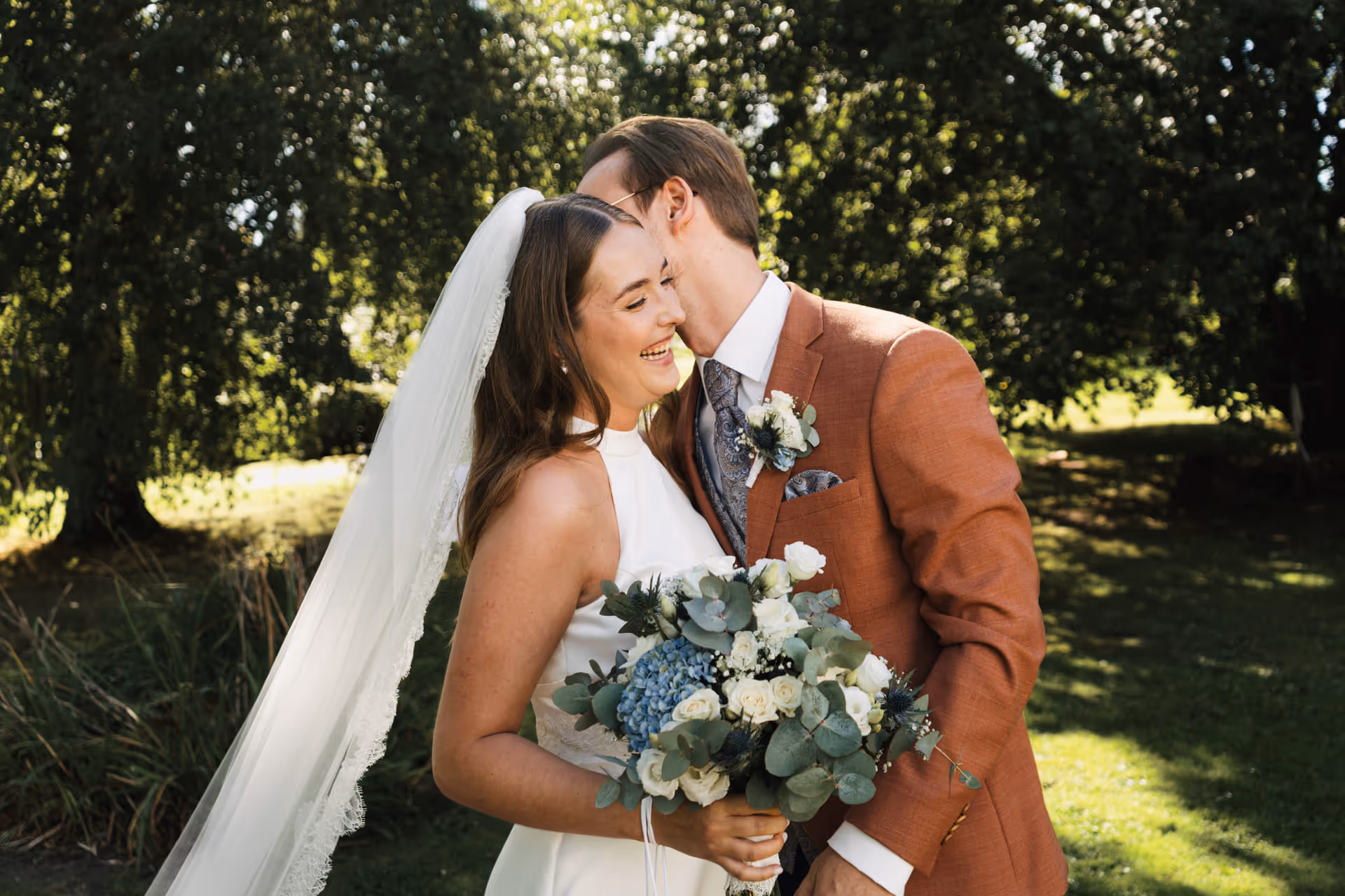 Bride in white dress with veil holding a bouquet, smiling as groom in brown suit leans in close outdoors.