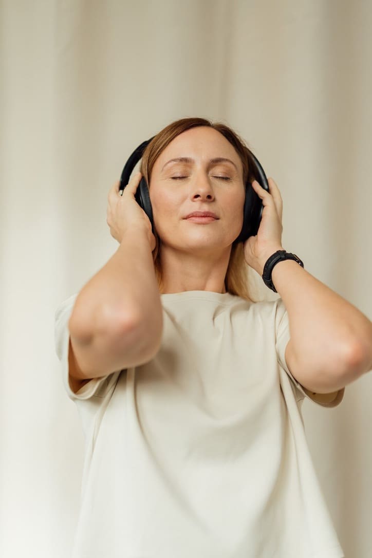 Woman with closed eyes enjoying music while wearing black headphones and a beige shirt.