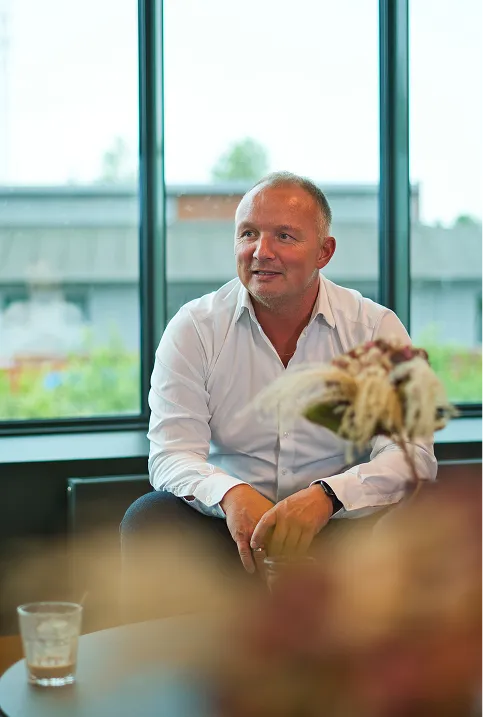 Smiling middle-aged man in a white shirt sitting indoors near large windows with a blurred bouquet and glass on the table in front.