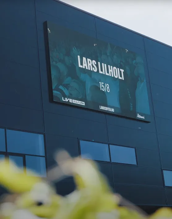Digital billboard on a dark building facade displaying 'LARS LILHOLT 15/8' with logos for Live Culture, Landsbyen.nu, and Albani.