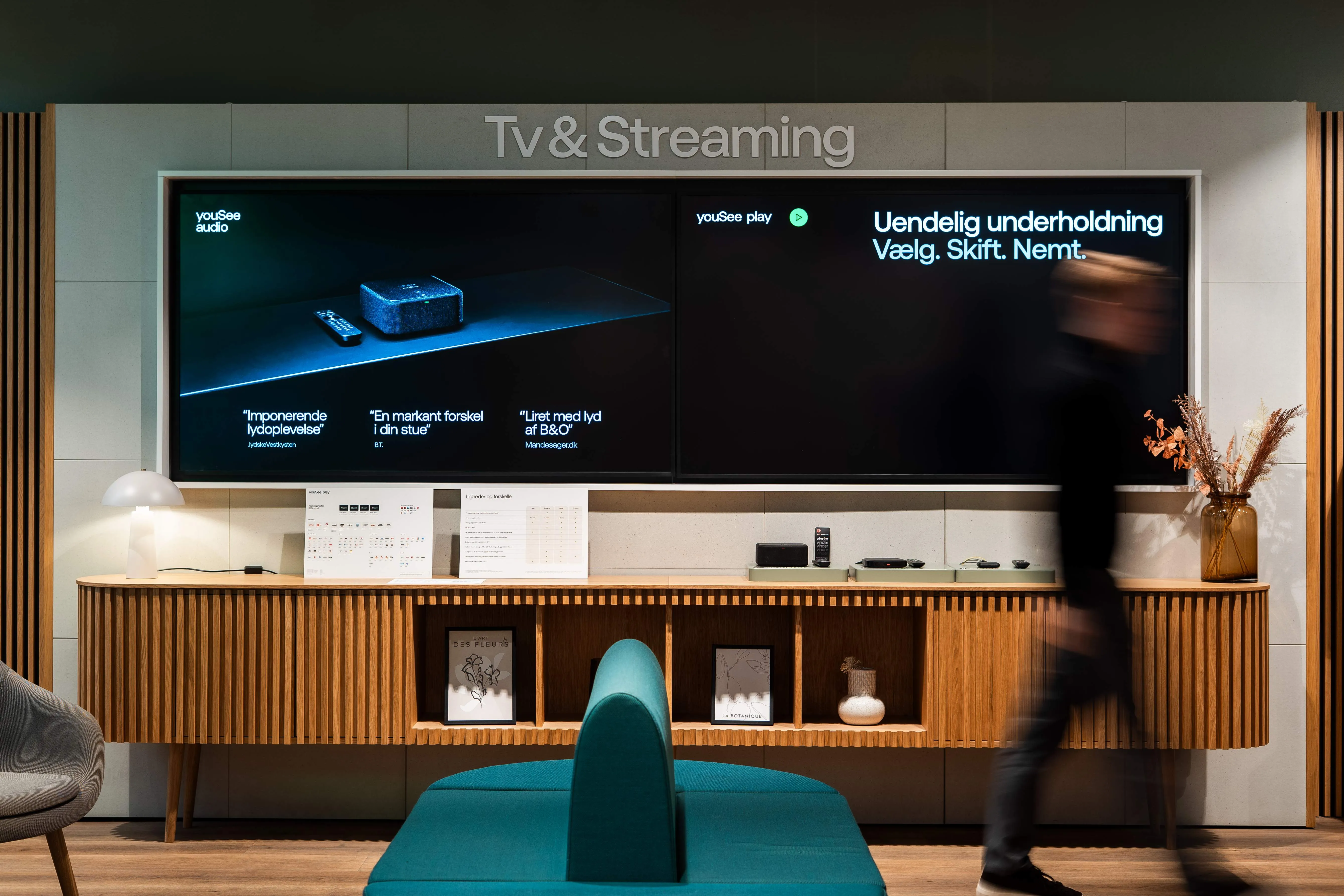 Modern showroom interior featuring a wooden console with tech devices, two large screens displaying Danish text about TV and streaming, and a blurred person walking past.