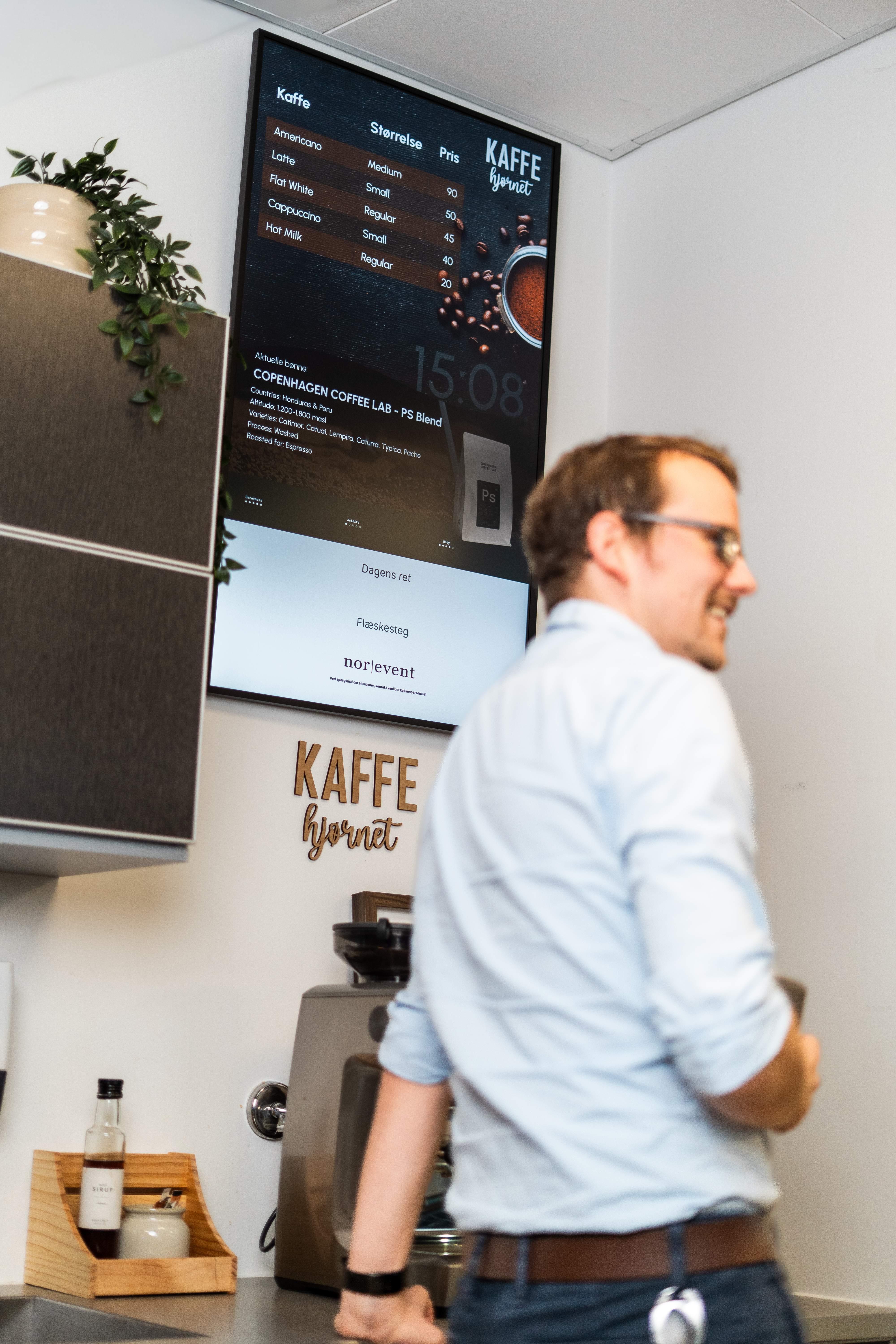 A man in glasses and a light blue shirt stands near a coffee station with a digital menu displaying coffee options and prices on the wall.