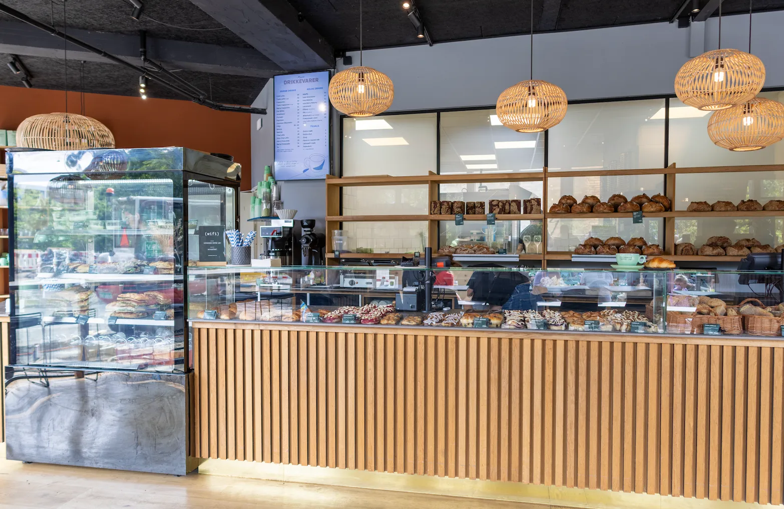 Modern bakery counter with wooden panels, glass display filled with assorted pastries and breads, and hanging wicker lamps above.