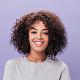 Smiling young woman with curly hair wearing a light gray sweater against a purple background.