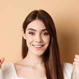 Smiling young woman with long straight brown hair wearing a white top against a beige background.