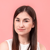 Portrait of a young woman with long brown hair wearing a white sleeveless top against a pink background.