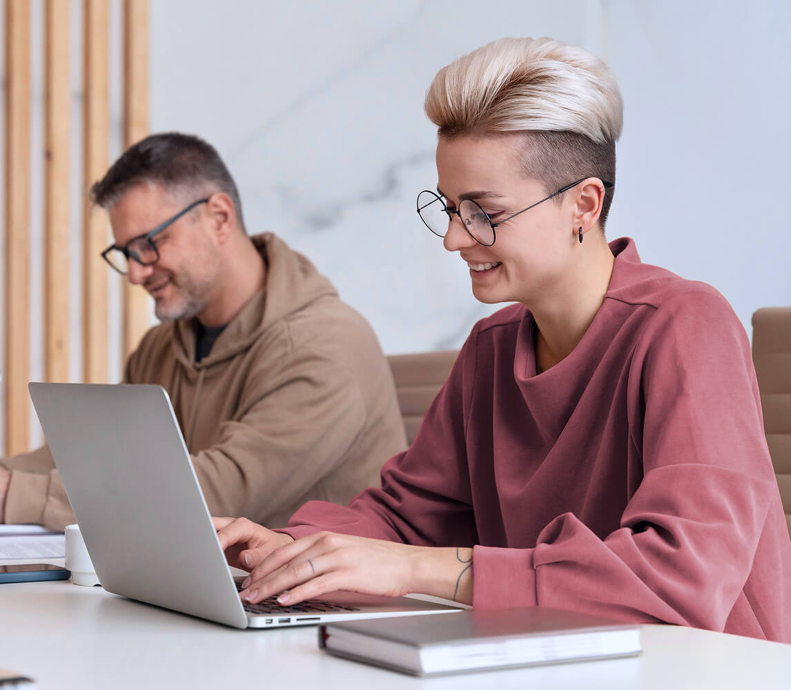 Two people working on laptops at a white desk in a modern office setting.