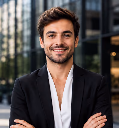 Young man with glasses, short brown hair, and a trimmed beard wearing a dark blazer and white shirt, standing outdoors with arms crossed.