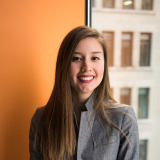 Smiling young woman with long brown hair wearing a gray jacket, standing indoors with orange and windowed background.