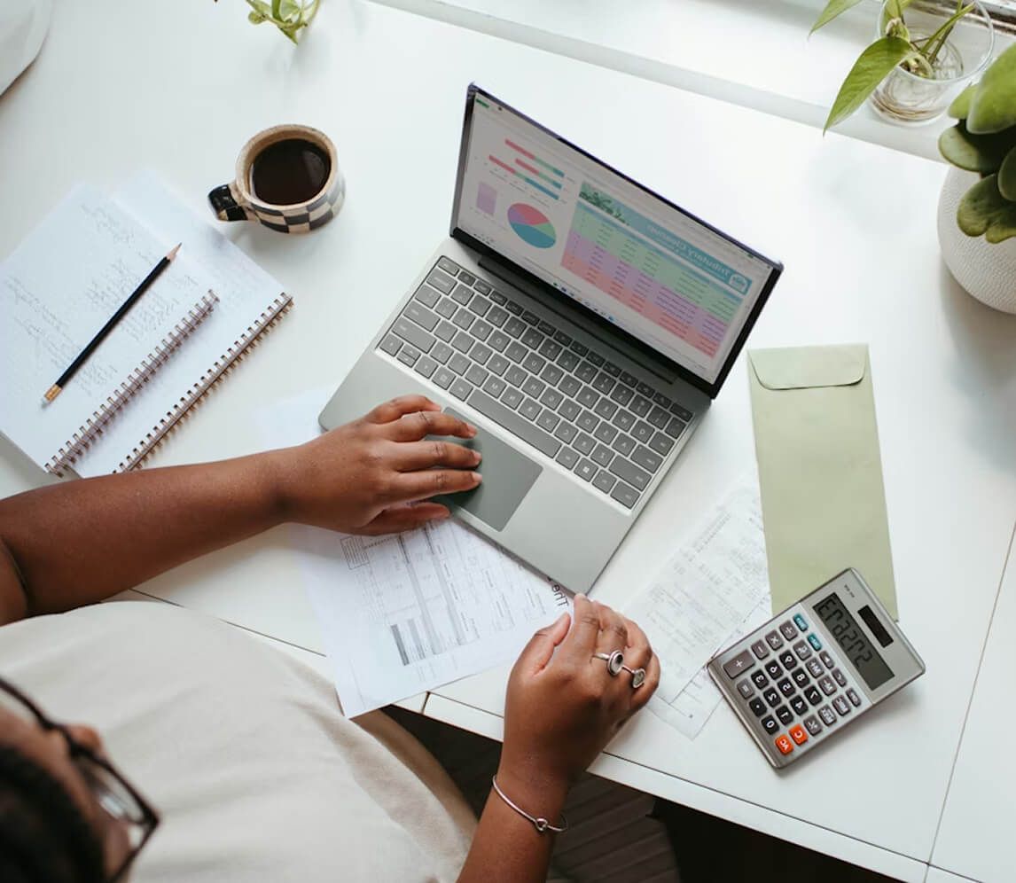Person working at a desk with a laptop displaying charts, a calculator, documents, a notebook, a pencil, a coffee cup, and potted plants.