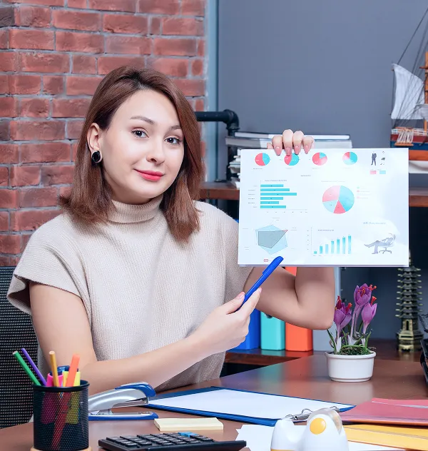 Young woman sitting at a desk in an office environment, holding up a sheet of paper with colorful charts and graphs and pointing at it with a blue pen.