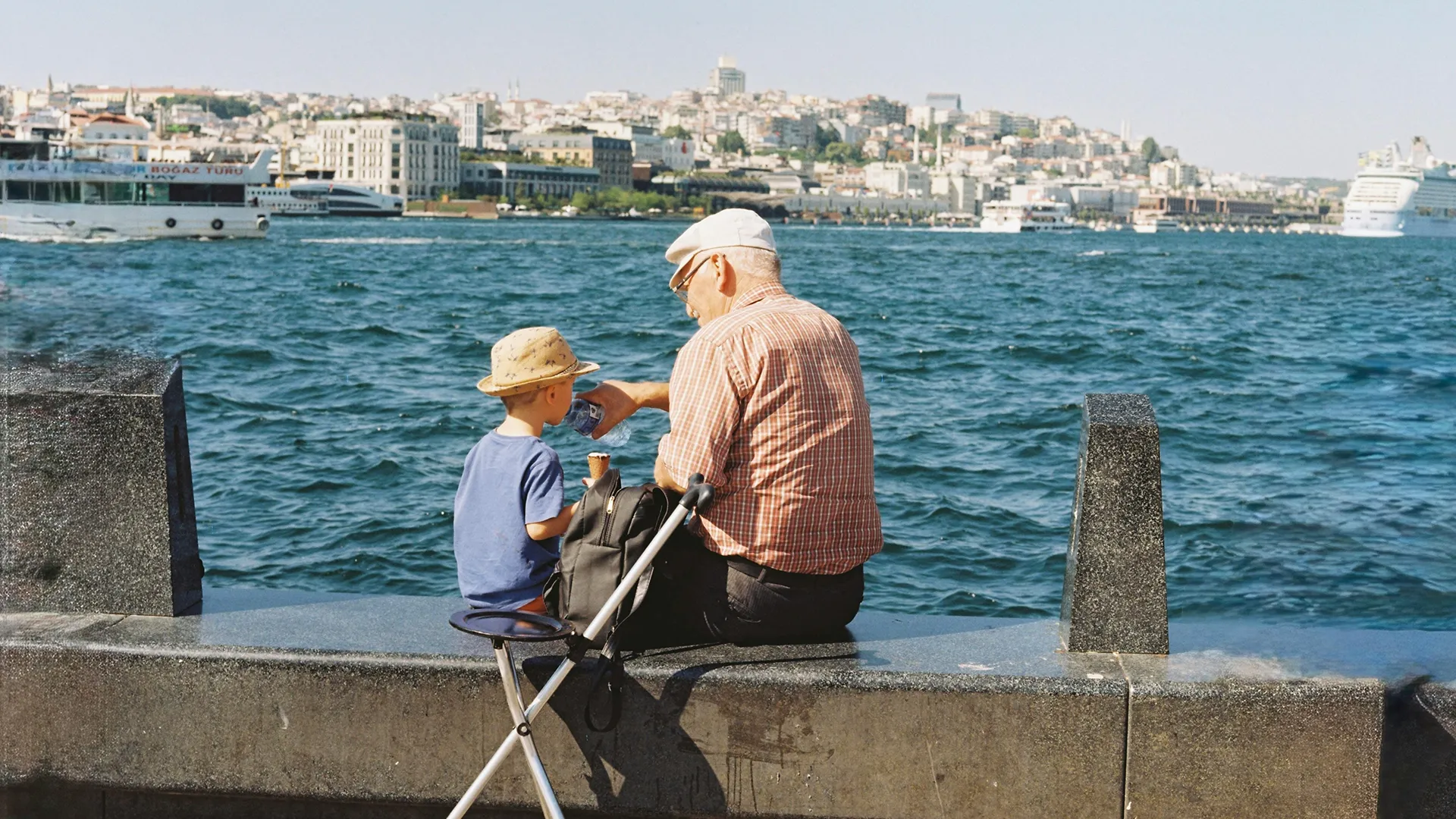 Oude man en jongen zitten samen aan de waterkant met ijsje en drinken, op de achtergrond een drukke stad en boten op het water.