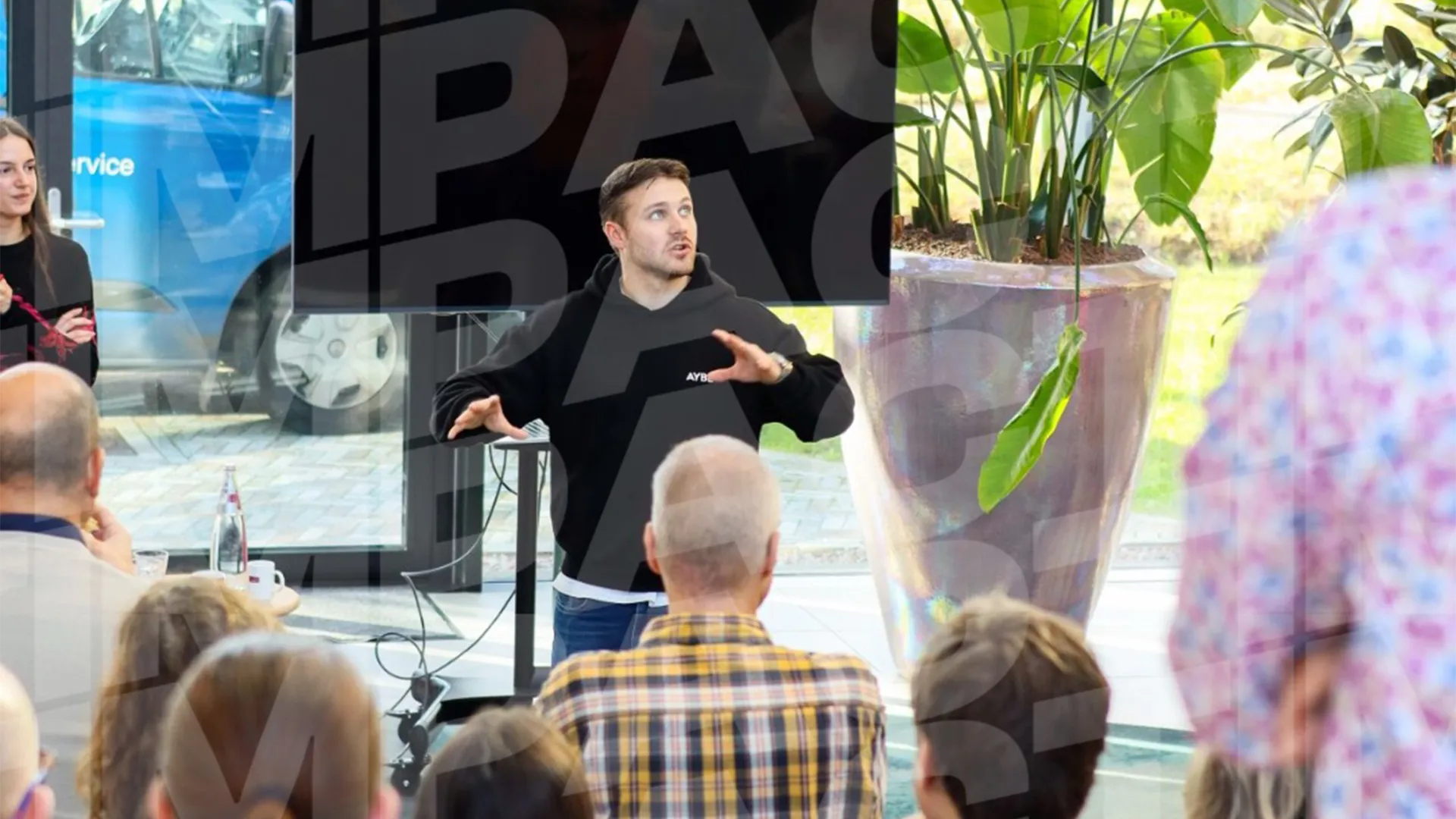 Man in zwarte hoodie geeft presentatie aan groep luisteraars in moderne ruimte met grote planten.