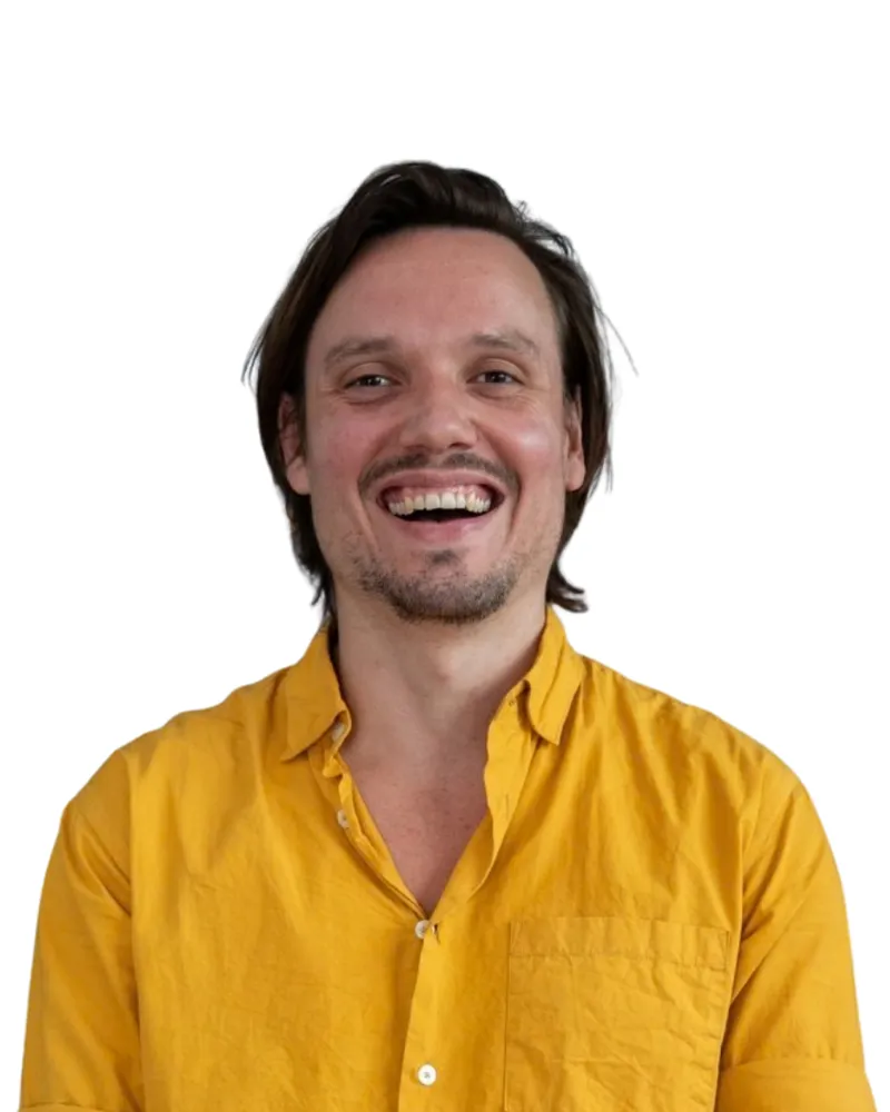 Man met een gele blouse lacht voor een achtergrond van bergen.