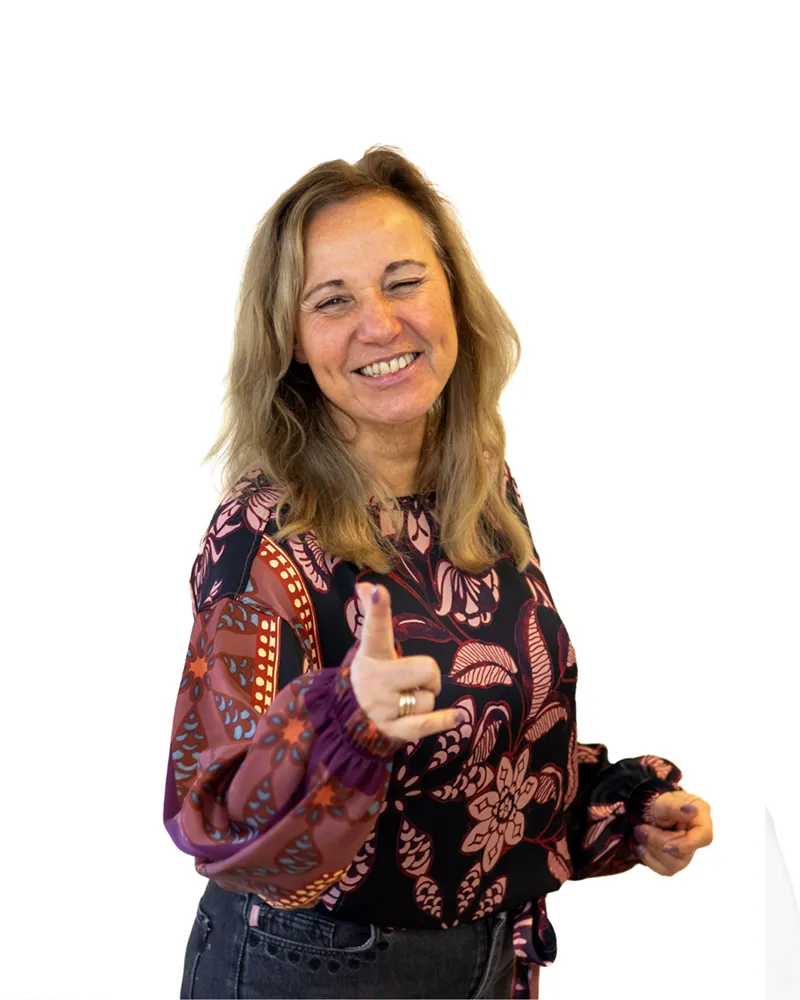 Vrouw in een kleurrijke blouse die glimlacht en naar de camera wijst.