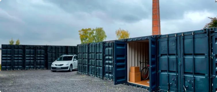 Série tmavě modrých kontejnerů na parkovišti za zatažené oblohy, jeden otevřený s viditelným kolem a krabicemi uvnitř, poblíž je bílý automobil.