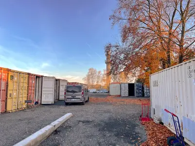 Venkovní sklad s nákladními kontejnery po obou stranách a stříbrným dodávkovým vozem parkovaným po levé straně, za jasného podzimního dne.