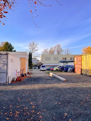 Podzimní průmyslové parkoviště s několika zaparkovanými auty mezi přenosnými kontejnery a budovami za jasného modrého nebe.