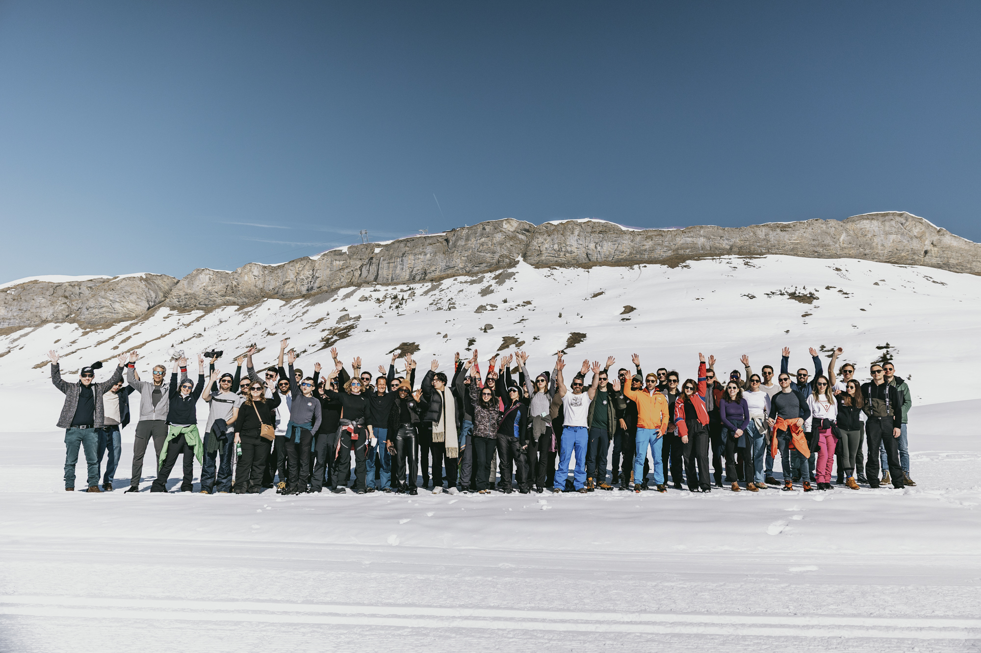 Large group of people standing in snowy mountain landscape waving under clear blue sky.