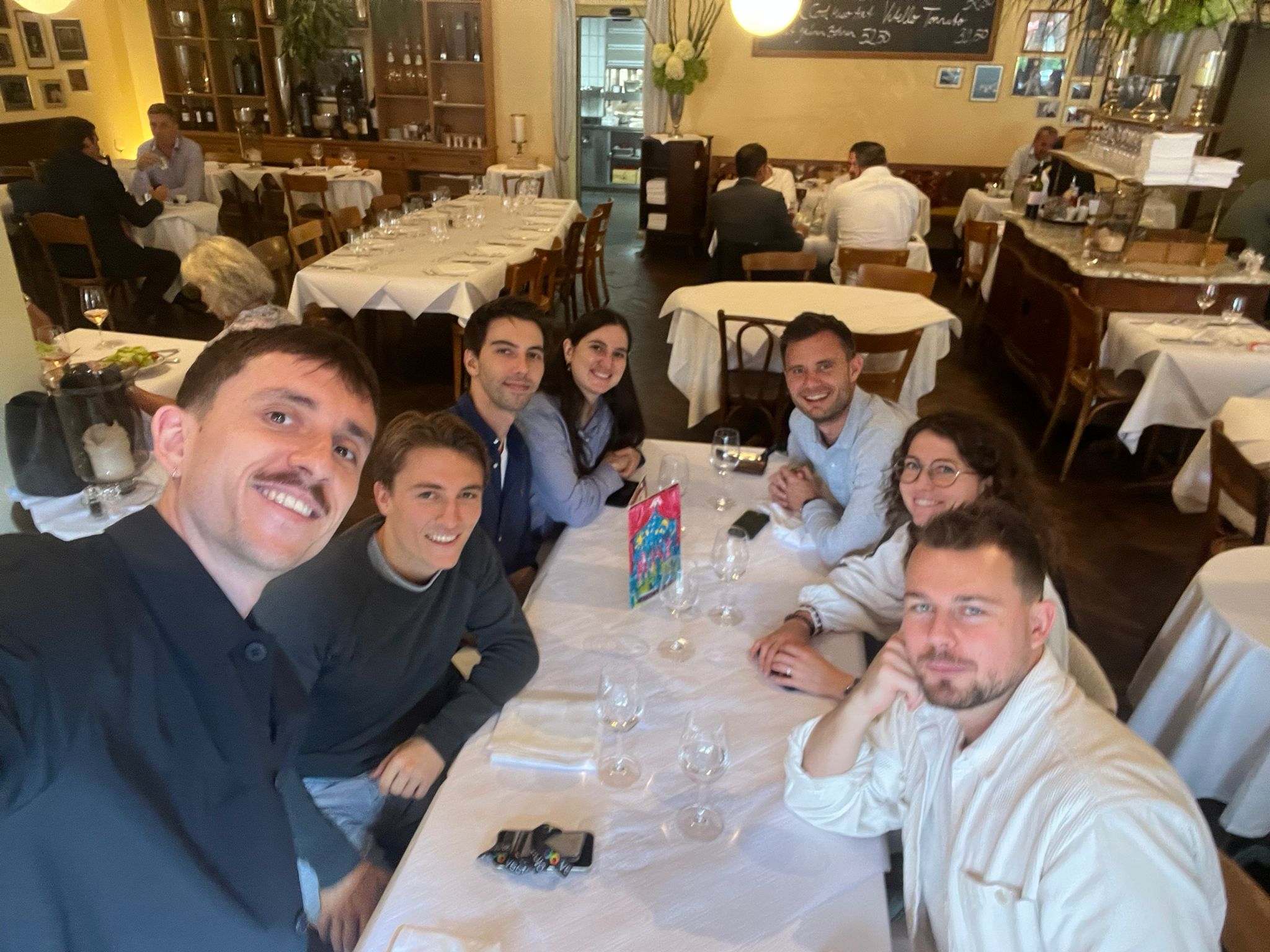 Seven people smiling at a restaurant table set with glasses and a colorful menu card.