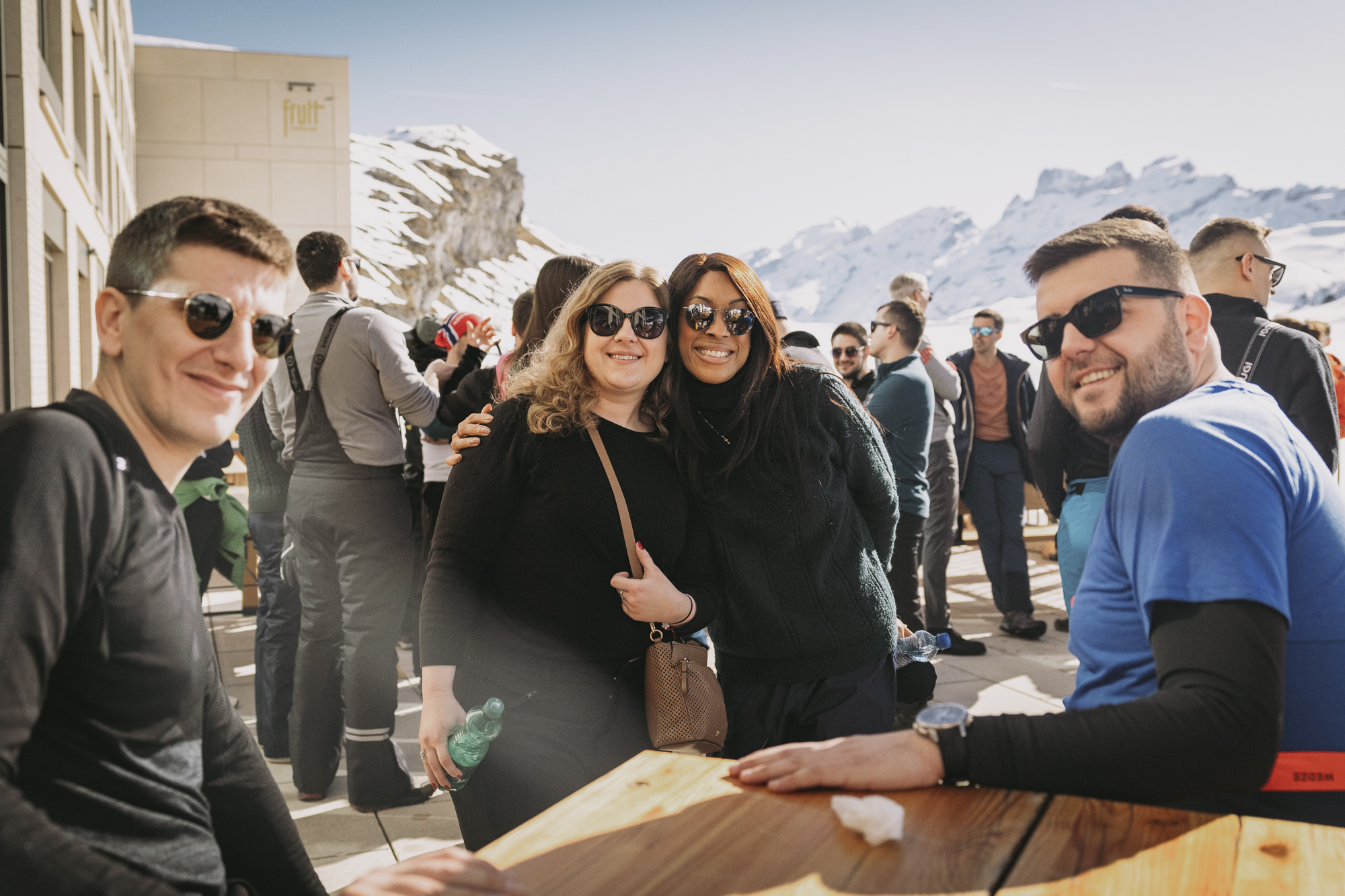 Group of friends wearing sunglasses outdoors with snowy mountain background on a sunny day.