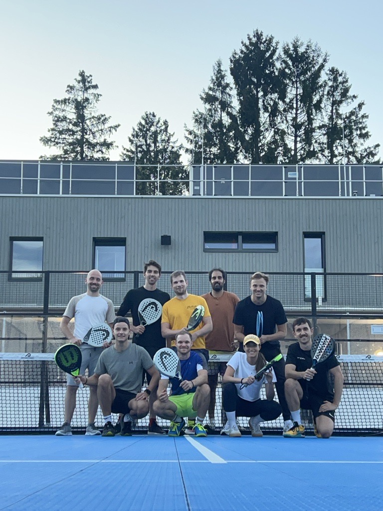 Group of nine people on a paddle tennis court posing with paddles in front of a building and trees.