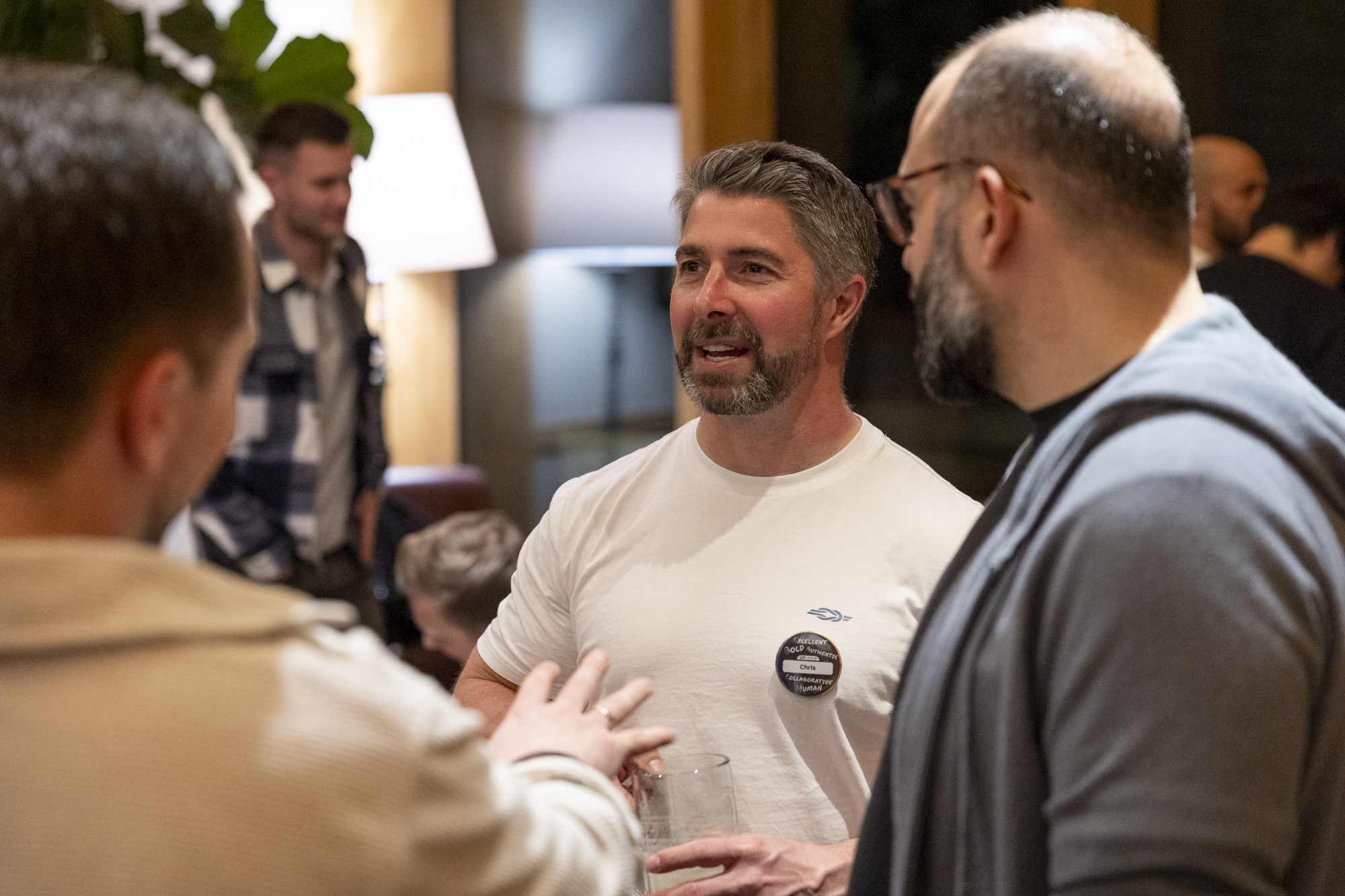 Three men standing and talking indoors, one man holds a glass, another wears glasses and a beard.
