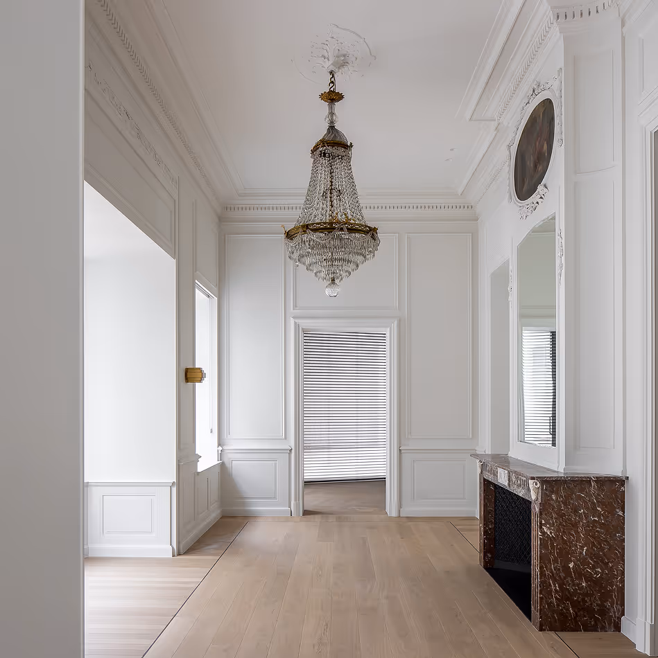 Minimalistische witte kamer met sierlijke kroonluchter, houten vloer, marmeren open haard en grote spiegel.