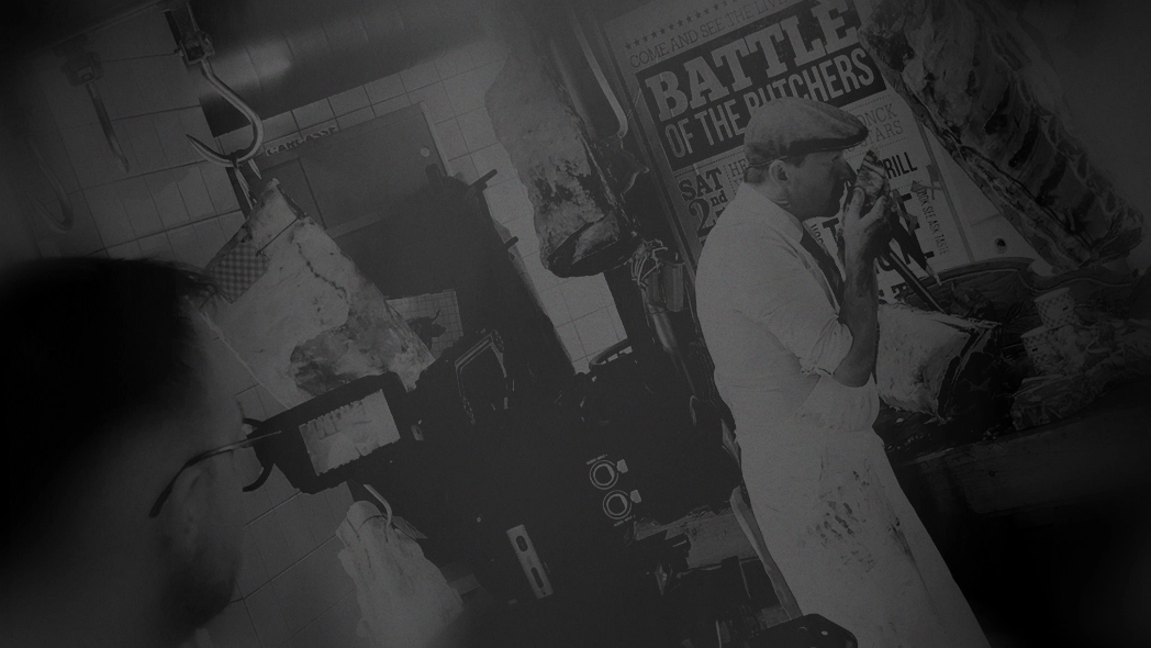 Butcher Hendrik Dierendonck in white apron and cap inspecting meat in a butcher shop with hanging cuts and a poster titled 'Battle of the Butchers'.
