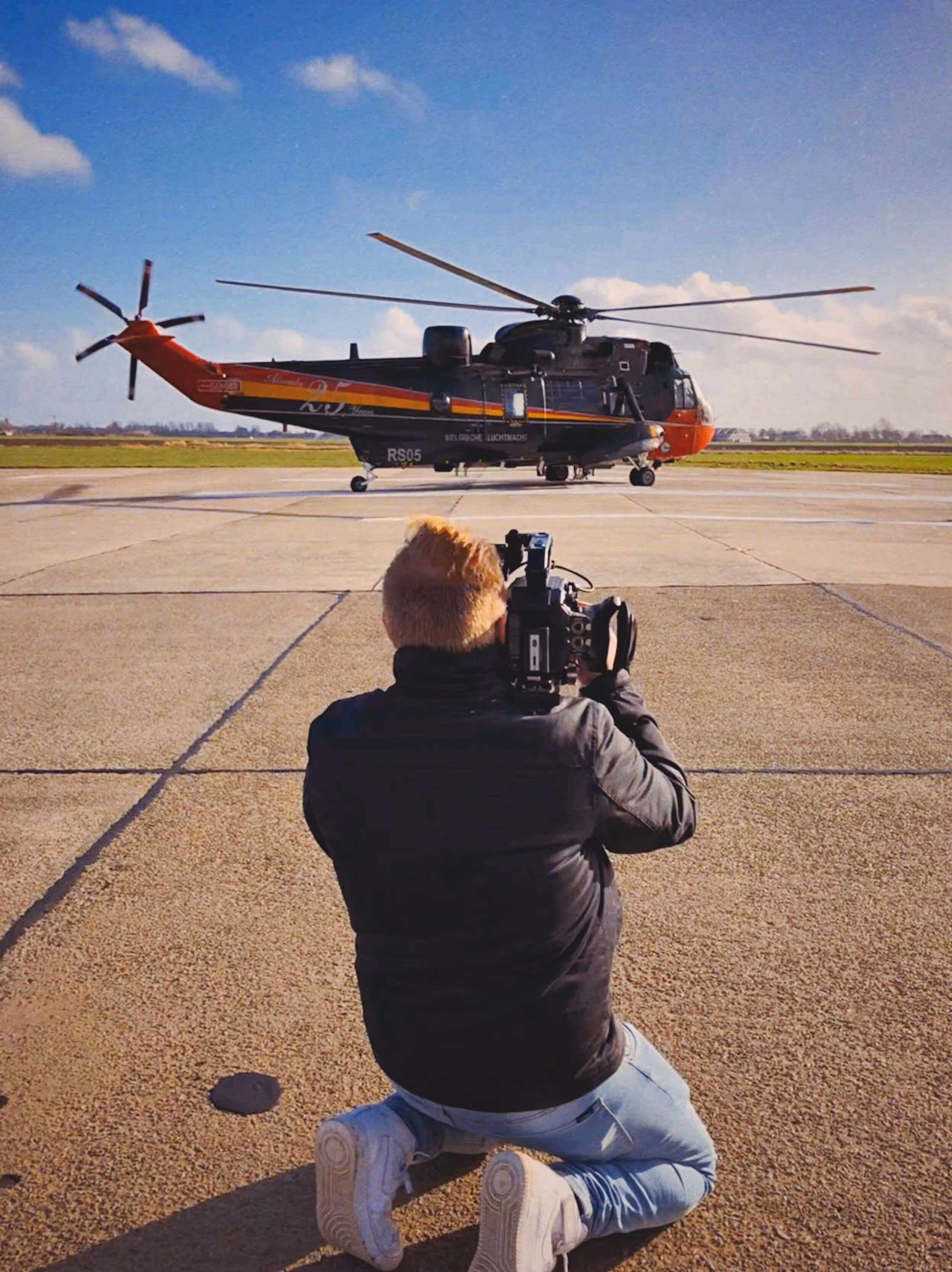 Person kneeling on a tarmac filming a black and orange helicopter (Seaking) with a video camera on a clear day.