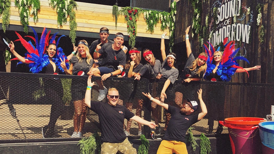 Group of smiling young adults posing behind a mesh fence with two wearing blue and red feathered costumes and others in casual black outfits with headbands, under greenery and a sign reading 'Sound of Tomorrow.'