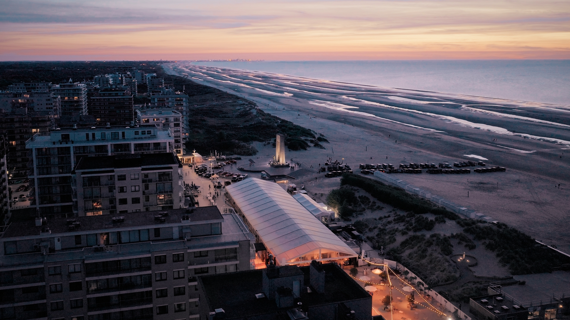 Aerial view of a coastal city at sunset with a long sandy beach in De Panne, buildings, a lit event tent, and parked cars along the shore.
