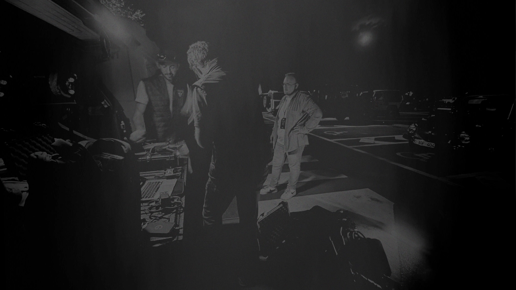 Three men standing around DJ equipment and a laptop at night in a parking lot with cars in the background.