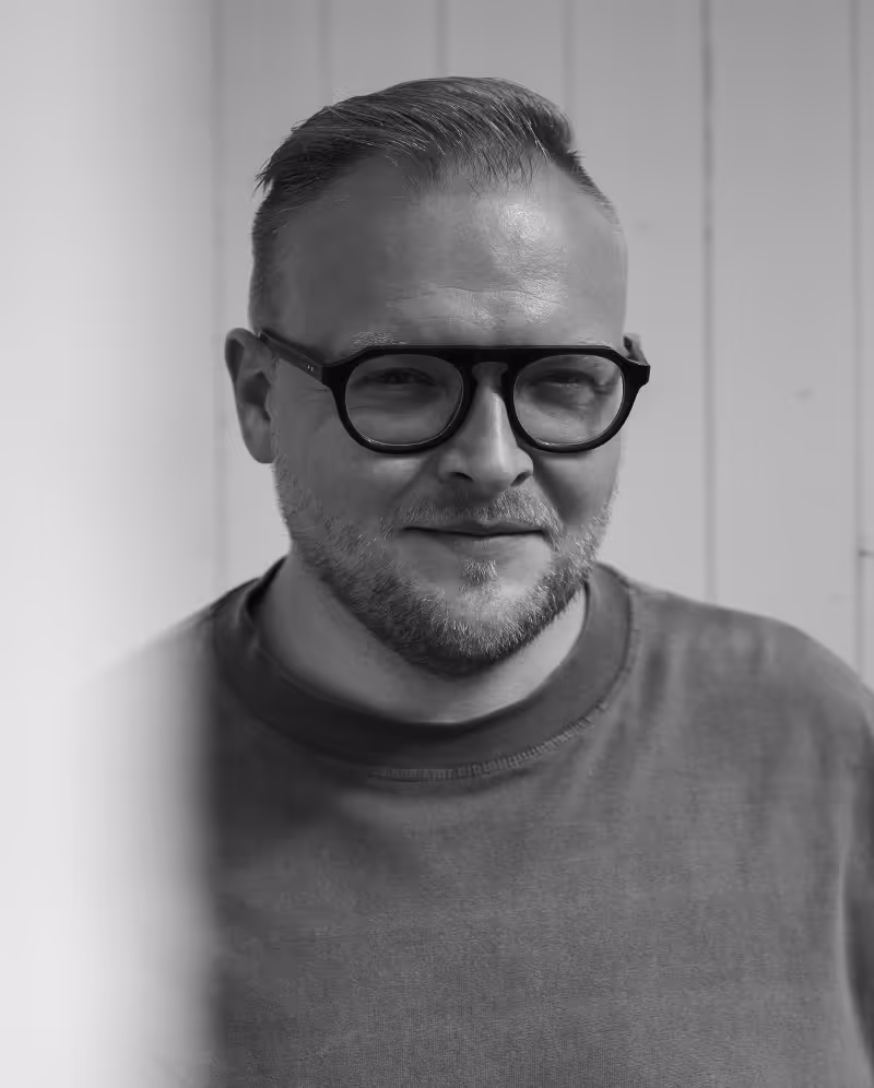 Man with short hair and beard wearing black-framed glasses and a crewneck shirt, smiling softly in front of a white paneled wall. Founder Nick Seys, Creative Agency Bypicknick.