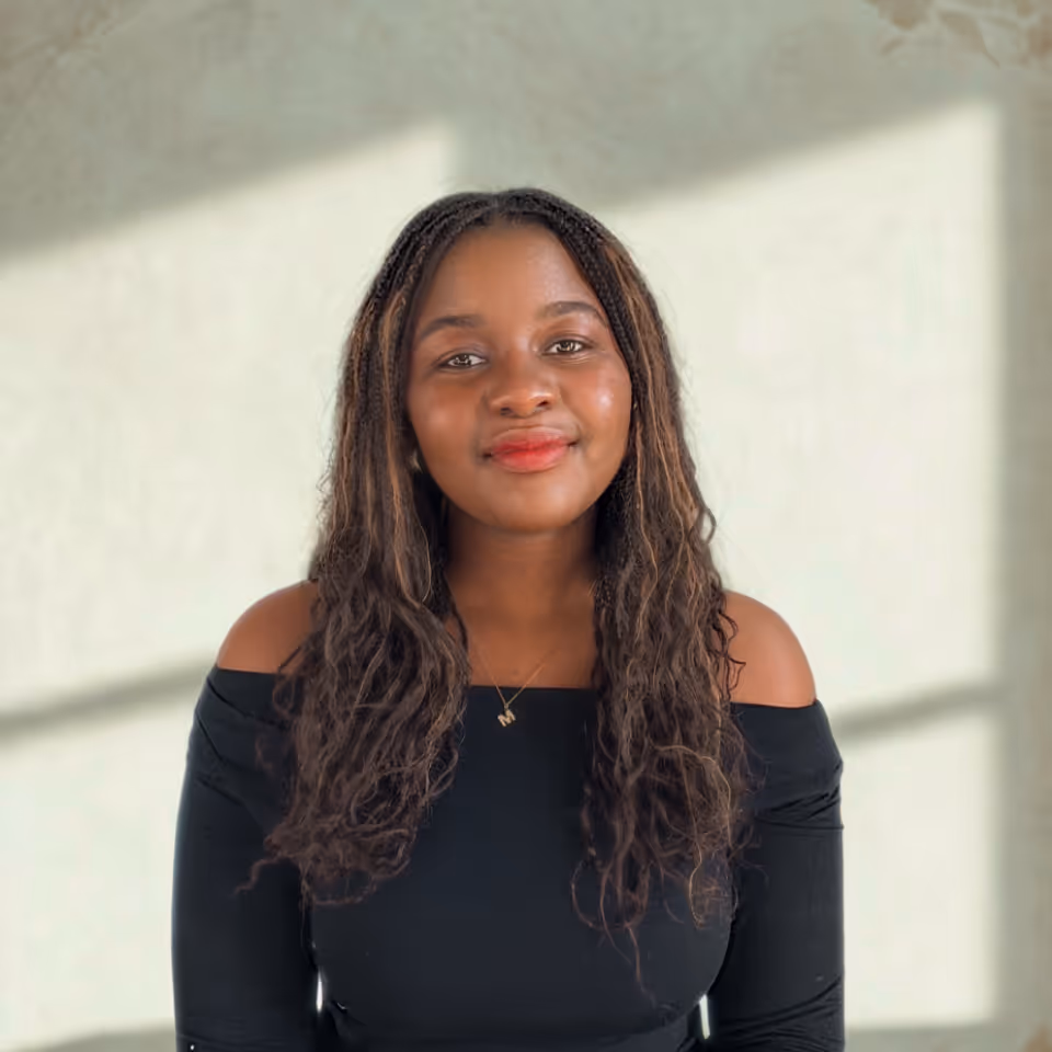 Smiling woman with long curly hair wearing an off-shoulder black top and a necklace with an 'M' pendant, standing against a light wall with window shadows.