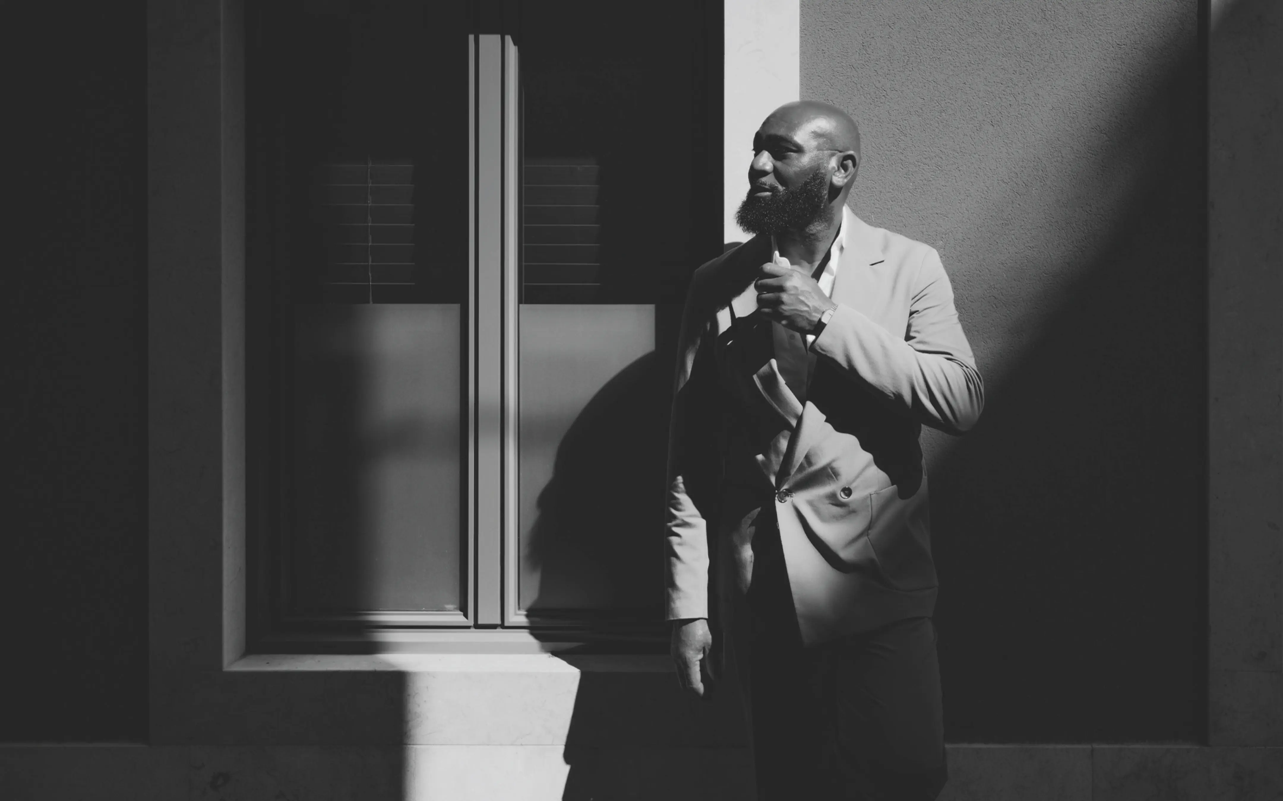 Black and white photo of a bearded man in a suit standing near a window with strong shadows on the wall.