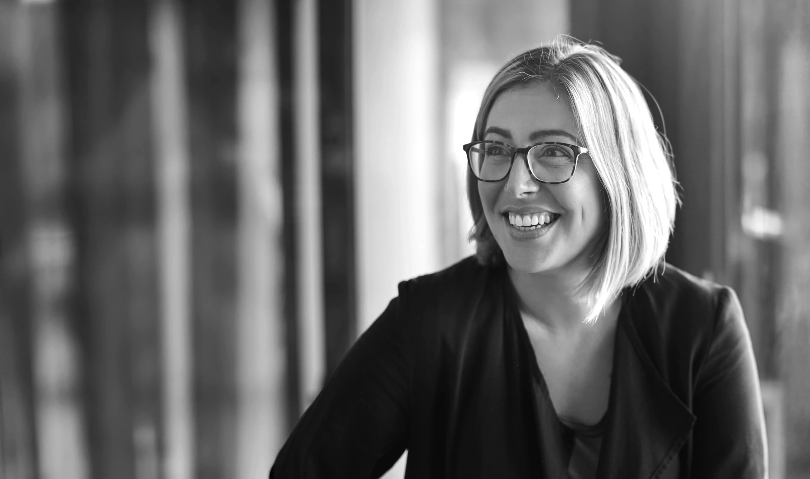 Smiling woman with glasses and shoulder-length hair wearing a dark jacket, looking to the side.